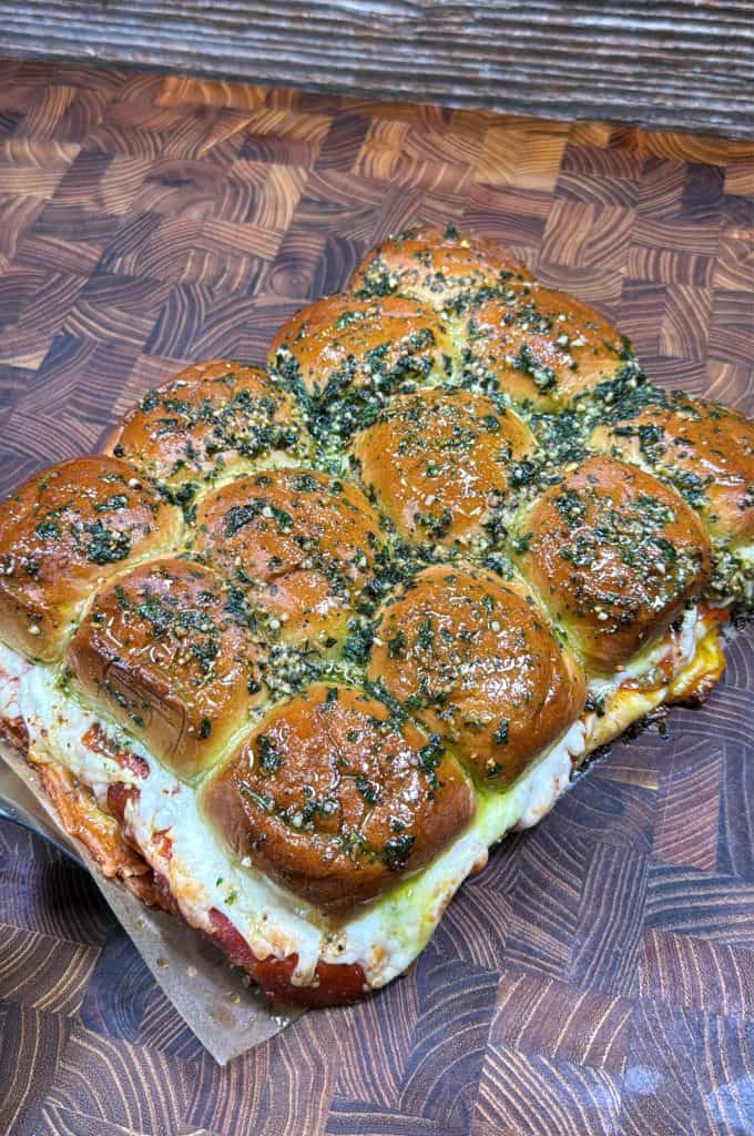 A batch of golden-brown, cheesy slider sandwiches topped with chopped herbs and garlic sits on a rustic wooden surface. The bread looks soft and the melted cheese is visible between the layers.