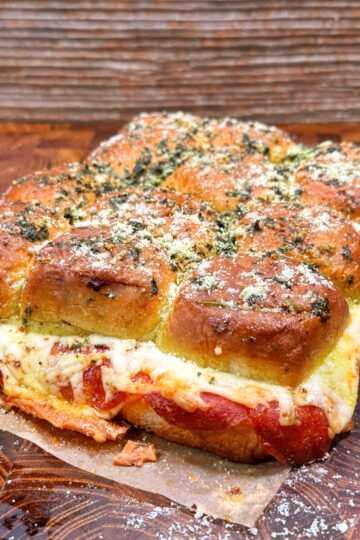 A batch of pull-apart sliders filled with melted cheese, sauce, and sprinkled with herbs and grated cheese, sits on parchment paper atop a wooden surface.