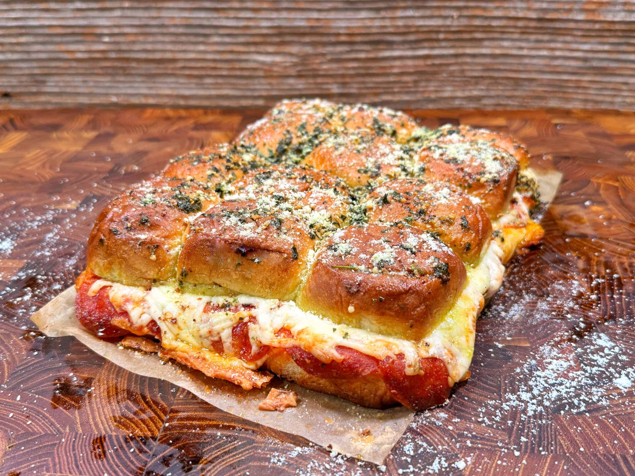 A batch of pull-apart sliders filled with melted cheese, sauce, and sprinkled with herbs and grated cheese, sits on parchment paper atop a wooden surface.