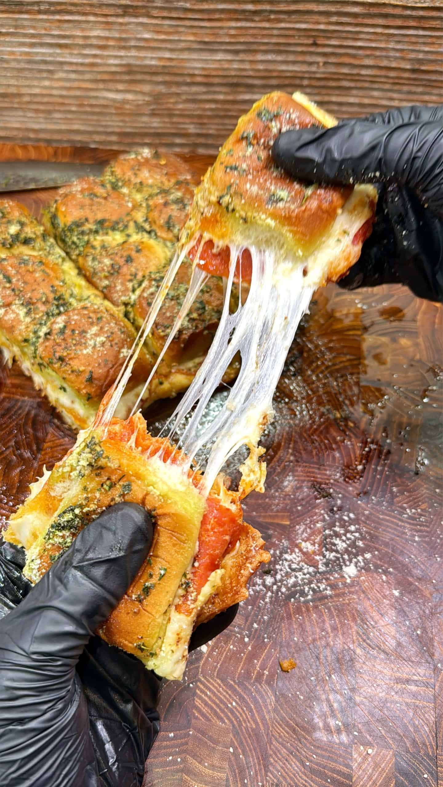 Two gloved hands pull apart a cheesy, herb-topped sandwich, showing melted cheese stretching between the halves. More pieces of the sandwich are visible on a wooden surface in the background.