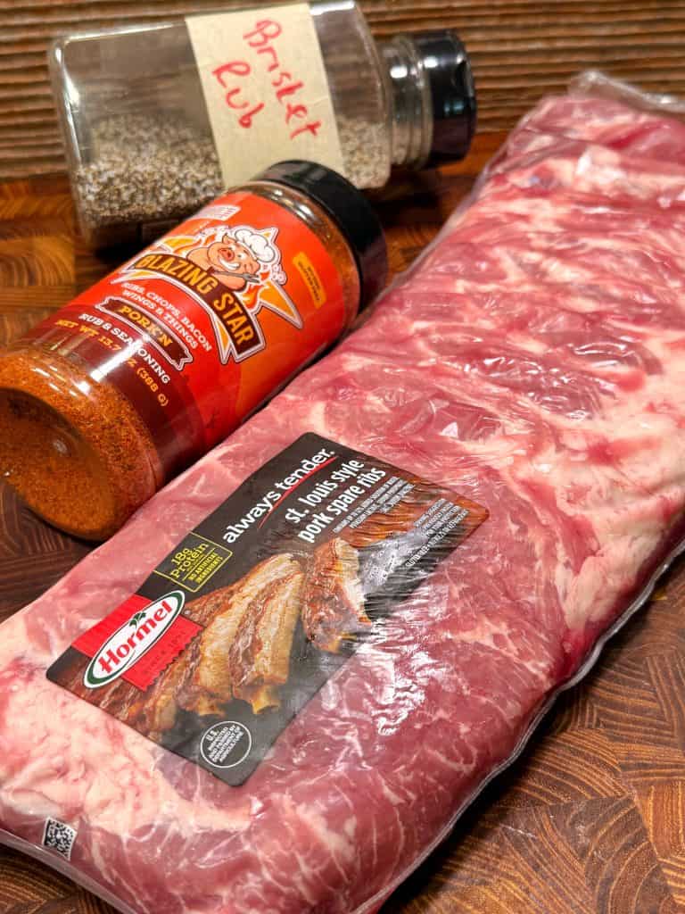 A package of Hormel St. Louis style pork spare ribs sits on a wooden surface next to a jar labeled Brisket Rub and a bottle of Blazing Star spicy dry rub seasoning.