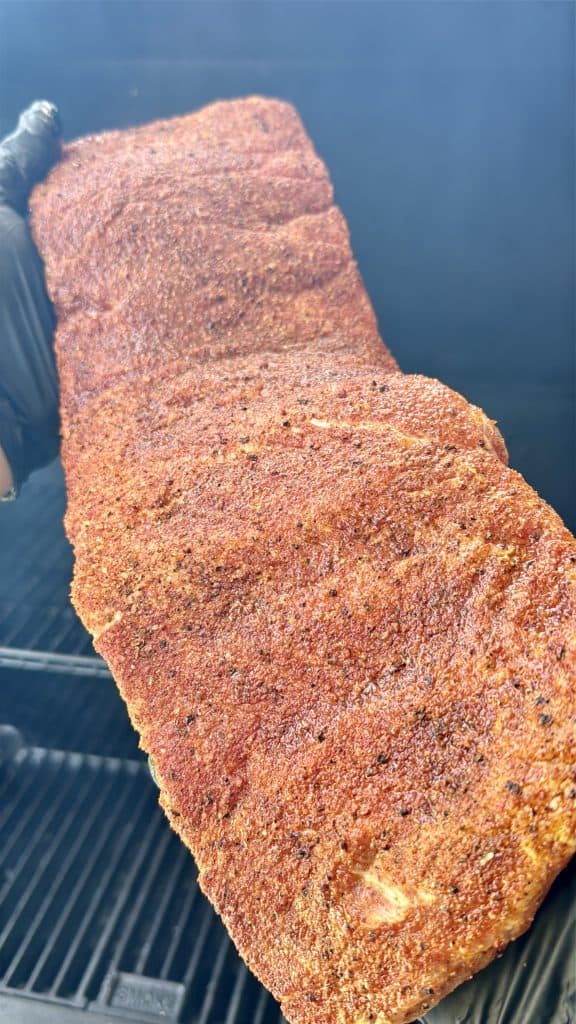 A gloved hand holds up a large, seasoned rack of ribs over a grill, ready to be smoked or cooked in a foil boat. The ribs are coated in a reddish-brown spice rub, with smoke visible in the background.