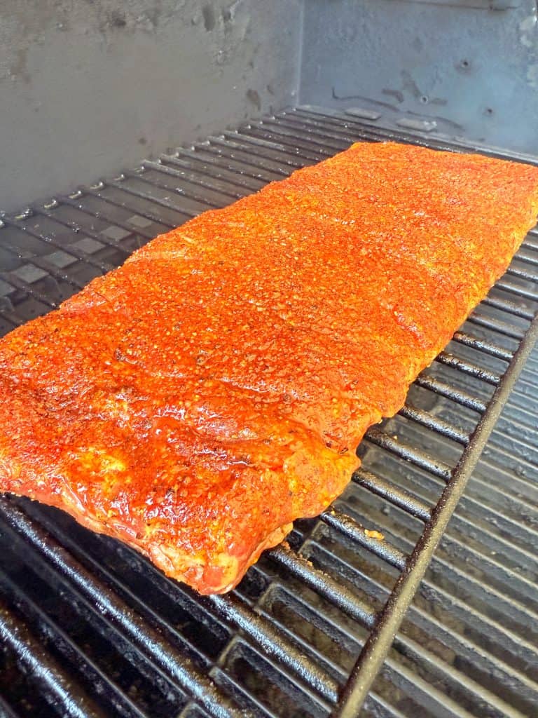 A large rack of ribs coated with reddish-brown seasoning is cooking on a grill inside a smoker. The meat appears uncooked and is placed on metal grates, with a dark interior background.