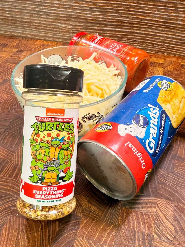 A bottle of Teenage Mutant Ninja Turtles Pizza Everything Seasoning, a can of Grands! biscuits, a jar of pizza sauce, and a bowl of shredded mozzarella cheese sit on a wooden surface.