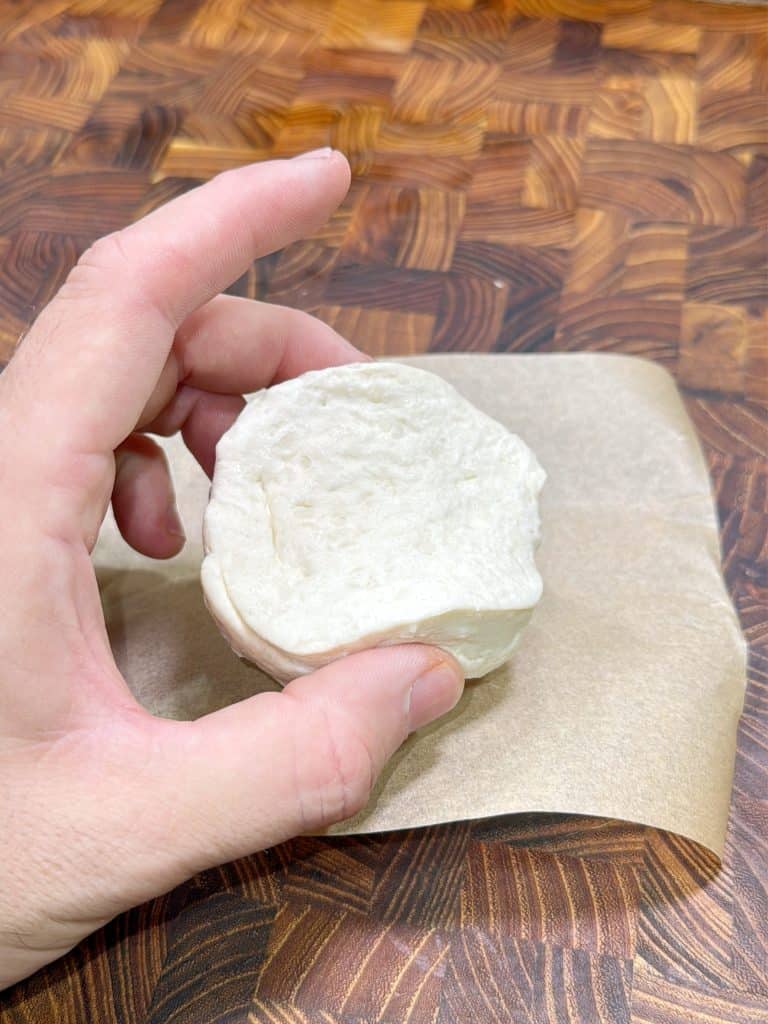 A hand holds an uncooked biscuit dough cup above a piece of parchment paper on a wooden patterned surface. The dough is shaped into a small, shallow cup.