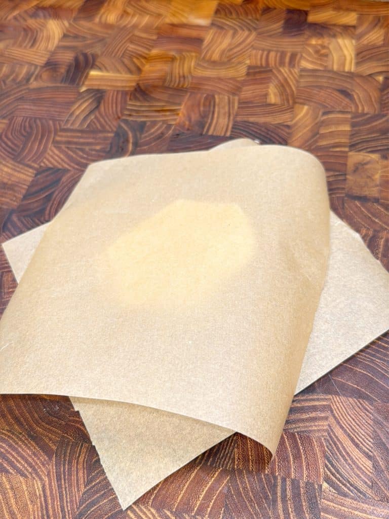 Two overlapping sheets of parchment paper, one with a translucent grease stain, placed on a wooden butcher block surface with a patterned design.