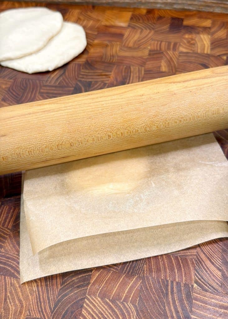 A rolling pin flattens dough between sheets of parchment paper on a wooden surface, with two rounds of dough visible in the background.