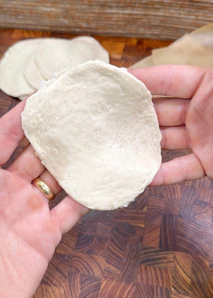 A person holding a piece of raw, flattened dough in their hands, with more pieces of dough on a wooden surface in the background.