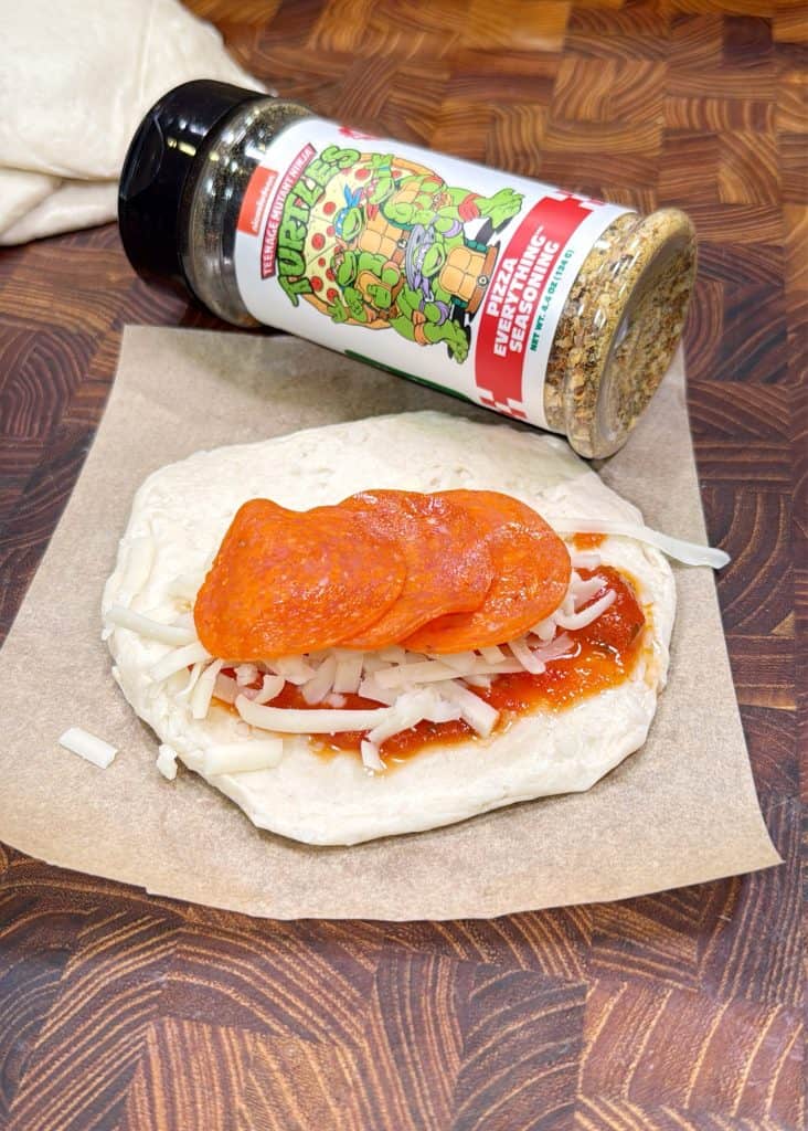 A piece of dough topped with pizza sauce, shredded cheese, and pepperoni slices sits on parchment paper. Behind it is a spice jar with Teenage Mutant Ninja Turtles branding that says Pizza Seasoning.