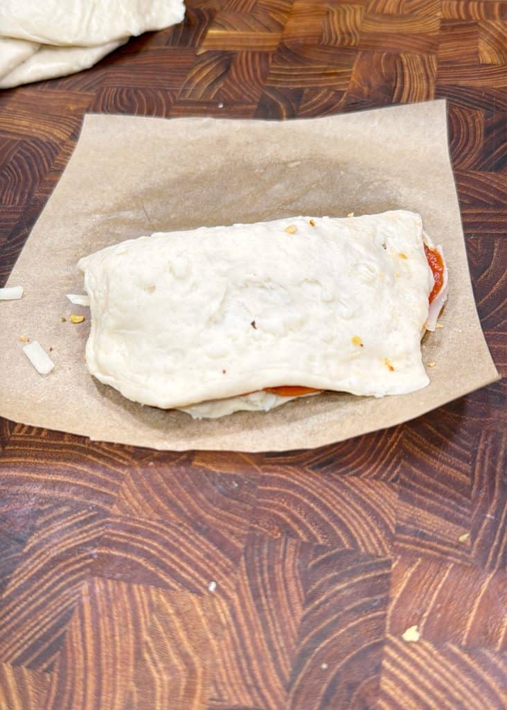 A folded, uncooked dough pocket with visible cheese and tomato inside sits on a piece of parchment paper atop a wooden cutting board to make biscuit calzones