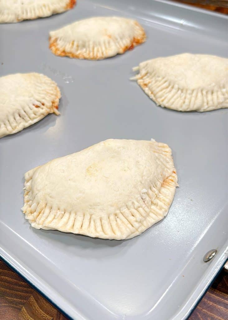 Unbaked hand pies with crimped edges on a light gray baking tray, ready to be cooked. The pastry dough looks soft and slightly floured.