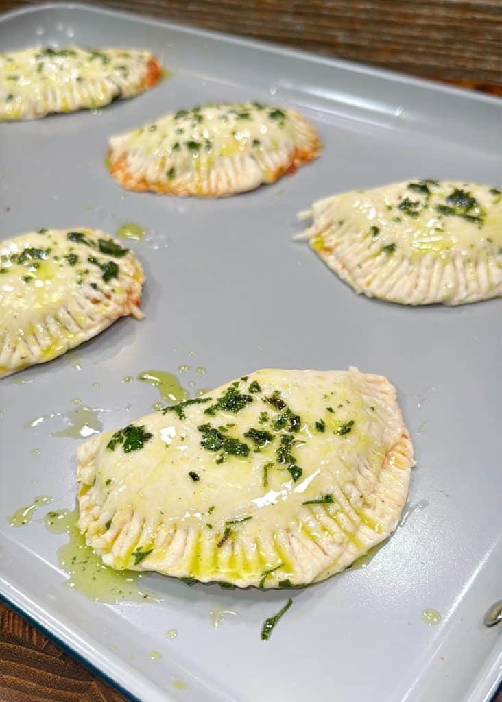 Six uncooked, half-moon shaped hand pies are arranged on a baking sheet. They are brushed with oil and sprinkled with chopped herbs, and have fork marks sealing the edges.