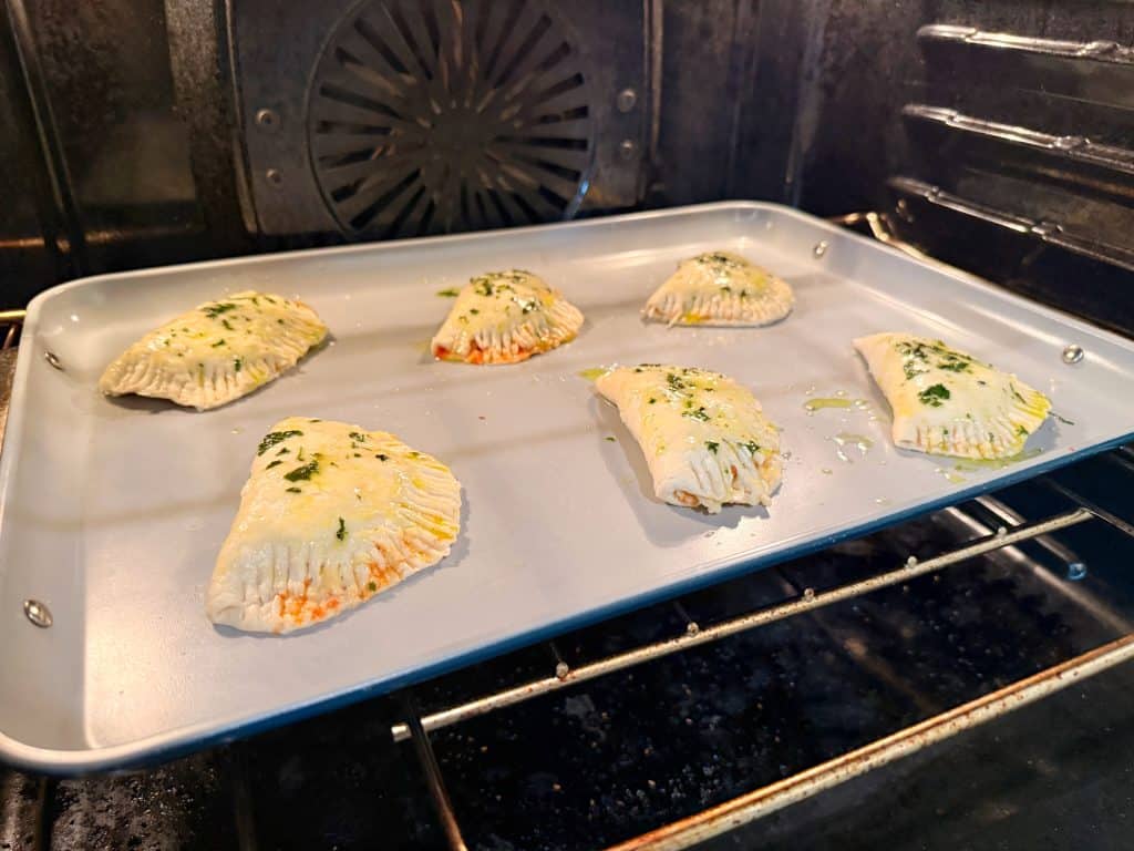 Six uncooked, herb-topped pastry turnovers are baking on a metal baking sheet inside an oven. The pastries have crimped edges and are spaced apart on the tray, with the oven rack and back visible.
