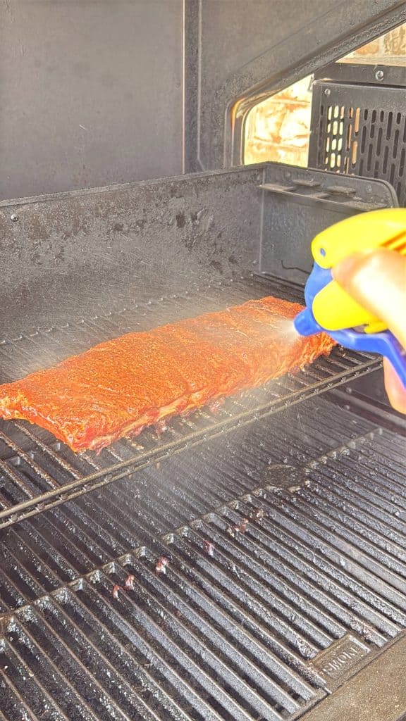 A rack of seasoned ribs on a grill is being misted with liquid from a yellow and blue spray bottle, likely to keep the meat moist while cooking.