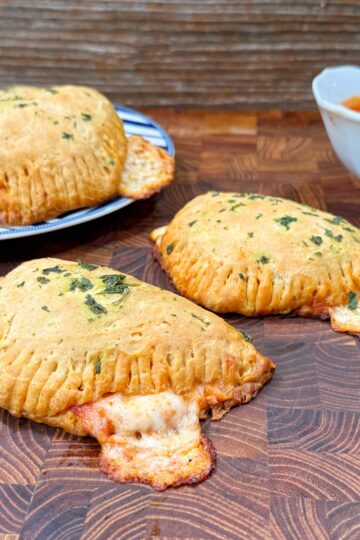 Three golden-brown mini calzones with melted cheese oozing out, garnished with herbs, are on a wooden surface. A bowl of marinara sauce and a plate with more calzones are in the background.