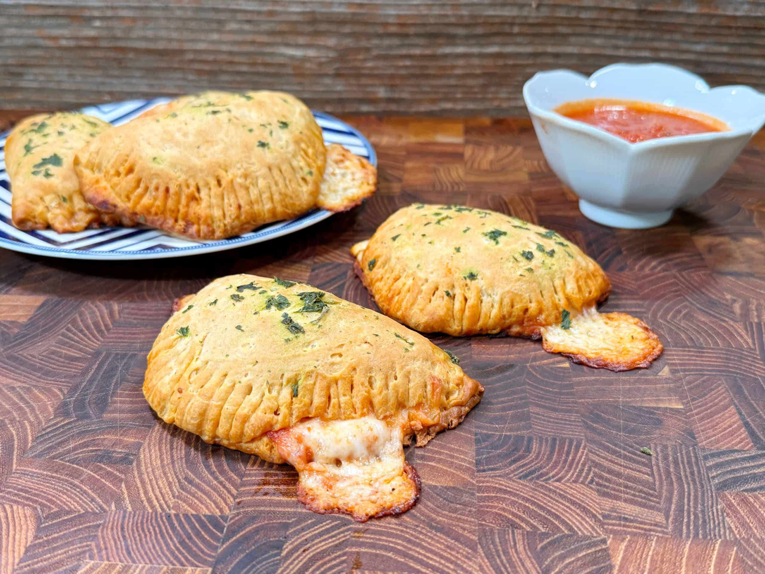 Three golden-brown mini calzones with melted cheese oozing out, garnished with herbs, are on a wooden surface. A bowl of marinara sauce and a plate with more calzones are in the background.