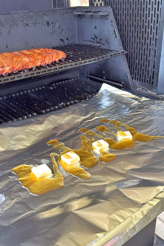 A barbecue grill with a rack of seasoned ribs on the upper grate. On the lower grate, a large sheet of foil holds several cubes of butter and drizzles of sauce or marinade.