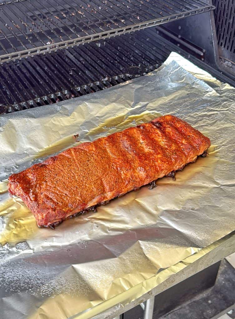 A rack of seasoned pork ribs sits on a sheet of aluminum foil on a grill, ready to be cooked. The grill grate is visible above the ribs.