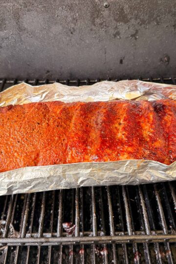 A rack of seasoned ribs covered in foil is cooking on a grill, with visible grill grates beneath and smoke marks on the meat.