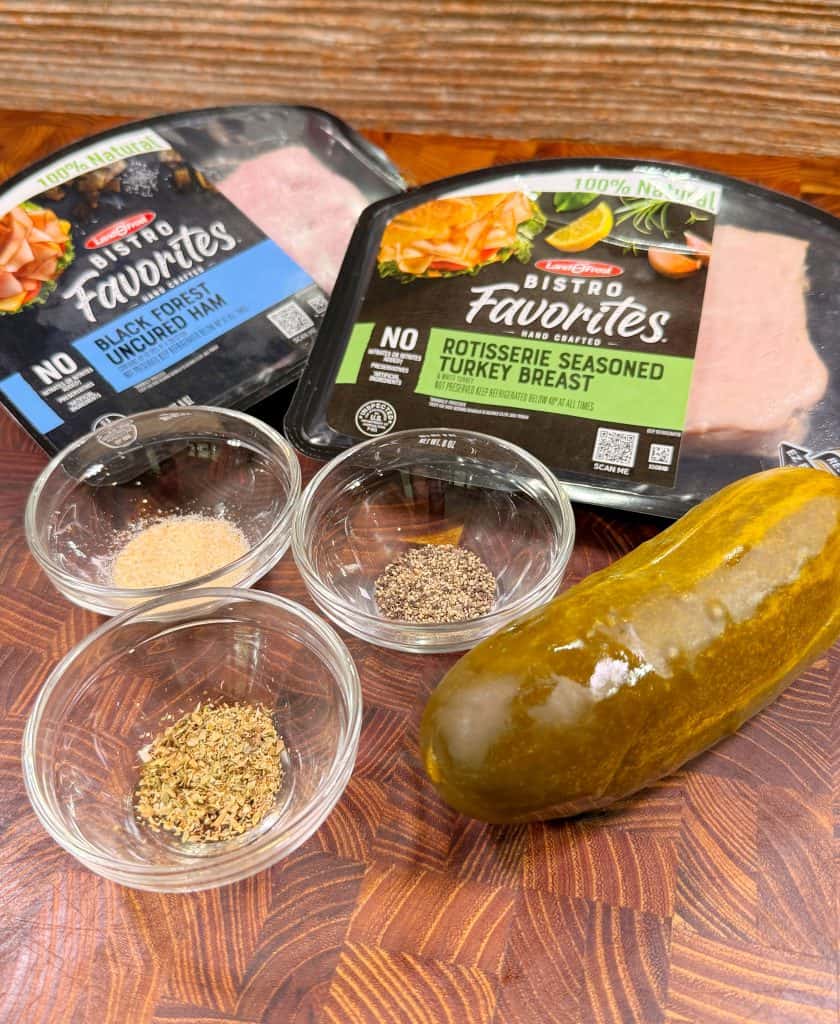 Two packages of lunch meat (black forest uncured ham and rotisserie seasoned turkey breast), a whole pickle, and three small bowls with various spices sit on a wooden surface.