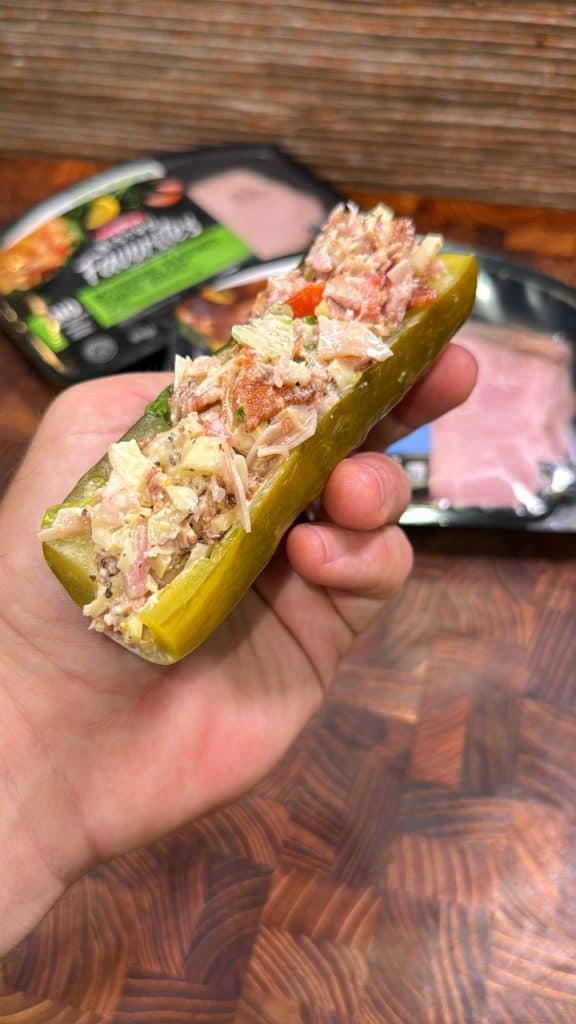 A hand holding half of a pickle stuffed with a creamy, mixed chopped turkey club that includes shredded meats and vegetables. Packaged deli meat is visible in the blurred background on a wooden surface.