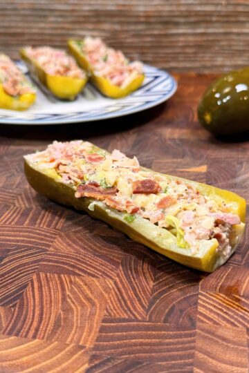 A pickle half stuffed with a creamy mixture sits on a wooden surface, with a whole pickle and a plate of more stuffed pickle halves in the background.