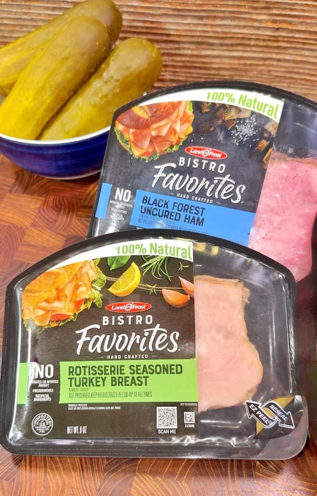 Two packs of Land OFrost Bistro Favorites deli meat—Black Forest Uncured Ham and Rotisserie Seasoned Turkey Breast—are on a table next to a blue bowl containing sliced pickles.