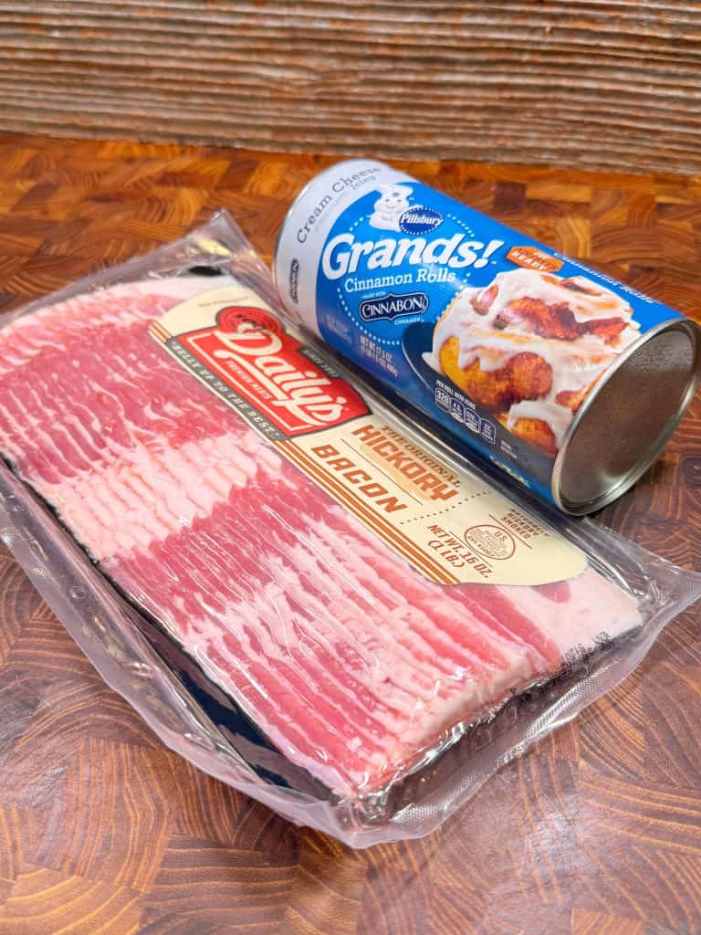 A package of hickory smoked bacon and a can of Pillsbury Grands! cinnamon rolls with cream cheese icing are placed side by side on a wooden surface.