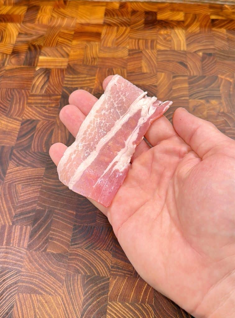 A hand holding two slices of raw bacon over a wooden cutting board with a checkered pattern.