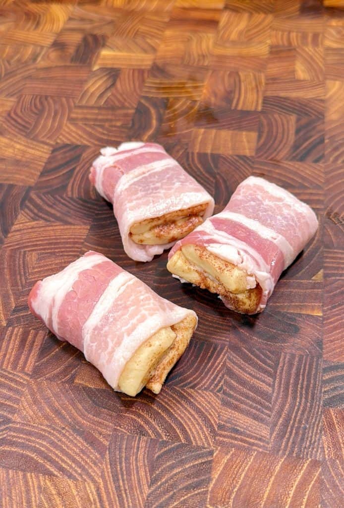 Three pieces of bacon-wrapped cinnamon roll bites are placed on a patterned wooden cutting board.