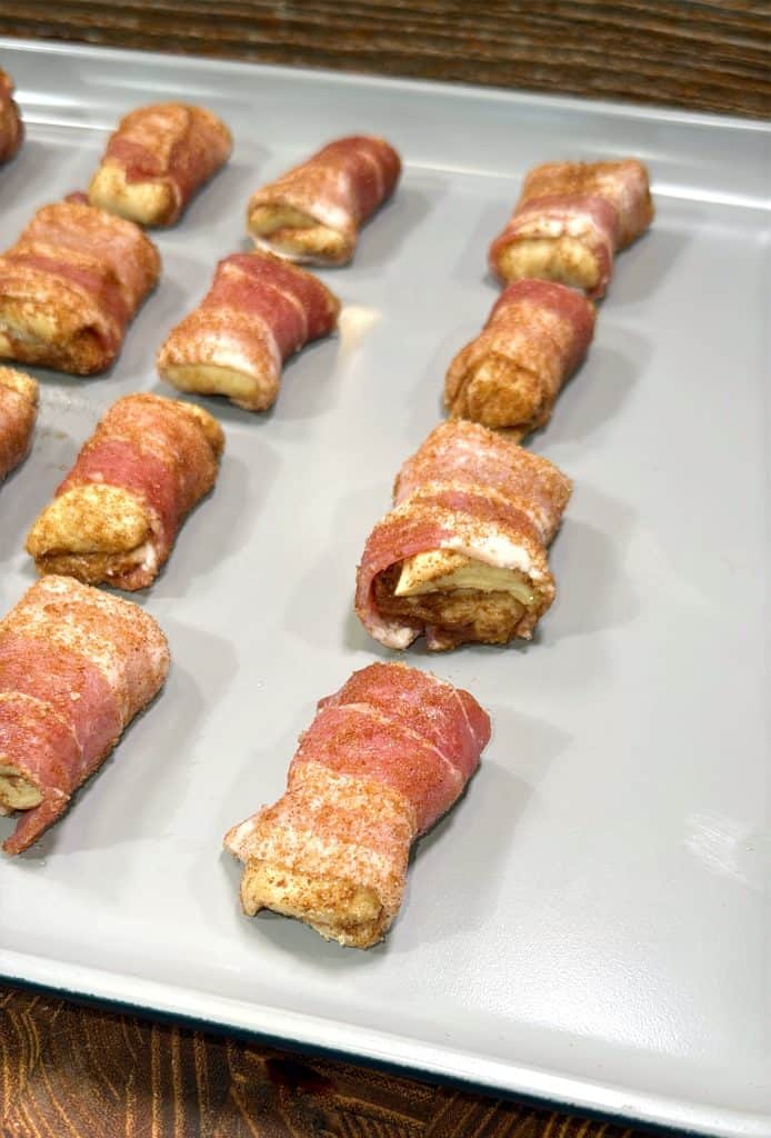 Bacon-wrapped, seasoned bites arranged in rows on a gray baking sheet, ready to be cooked. The surface beneath the sheet is a dark wood pattern.