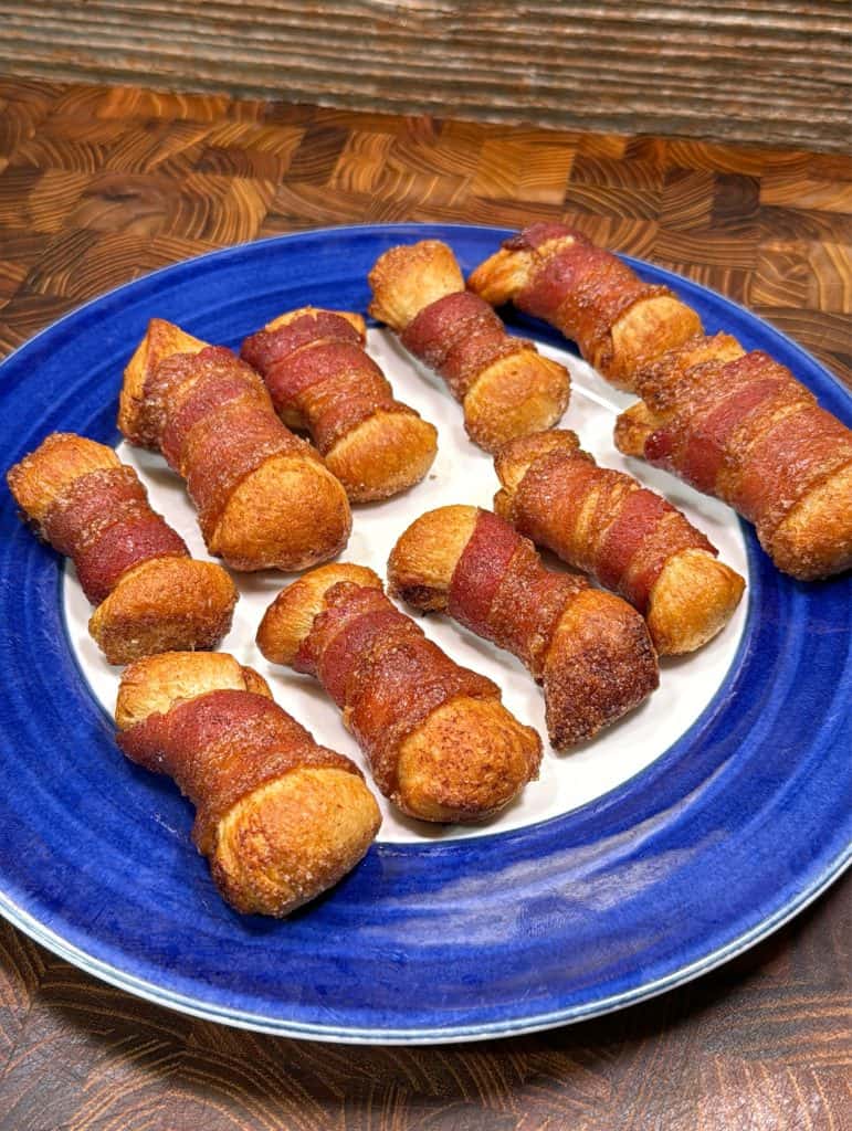 Eight bone-shaped pastries wrapped in bacon are arranged on a blue and white plate. The pastries are golden brown and sit on a wooden surface with a rustic background.