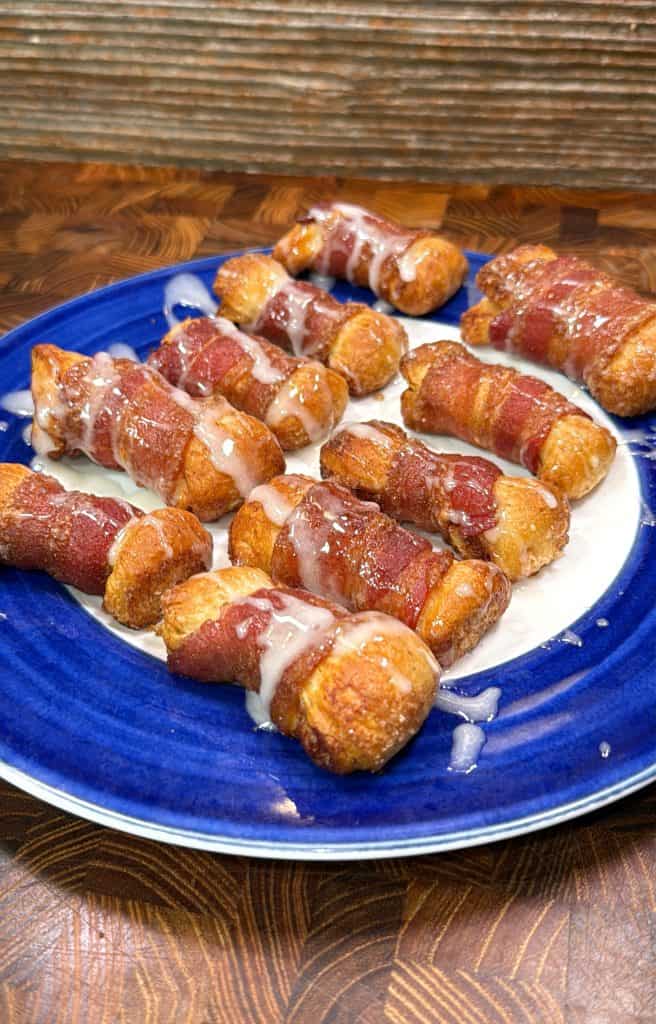 A blue plate holds several bacon-wrapped pastry twists drizzled with a white glaze, arranged on a wooden surface with a textured, brown background.