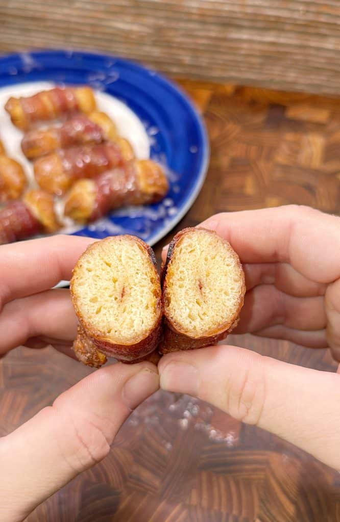 Two hands hold and split open a bacon-wrapped pastry, revealing its golden, airy inside. In the background, more bacon-wrapped pastries are served on a blue plate.