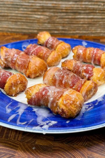 A blue plate holds several bacon-wrapped sausages drizzled with a creamy white sauce, set on a wooden surface with a textured wood background.