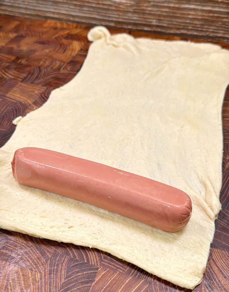 A raw hot dog rests on a rectangular sheet of uncooked dough placed on a wooden surface.