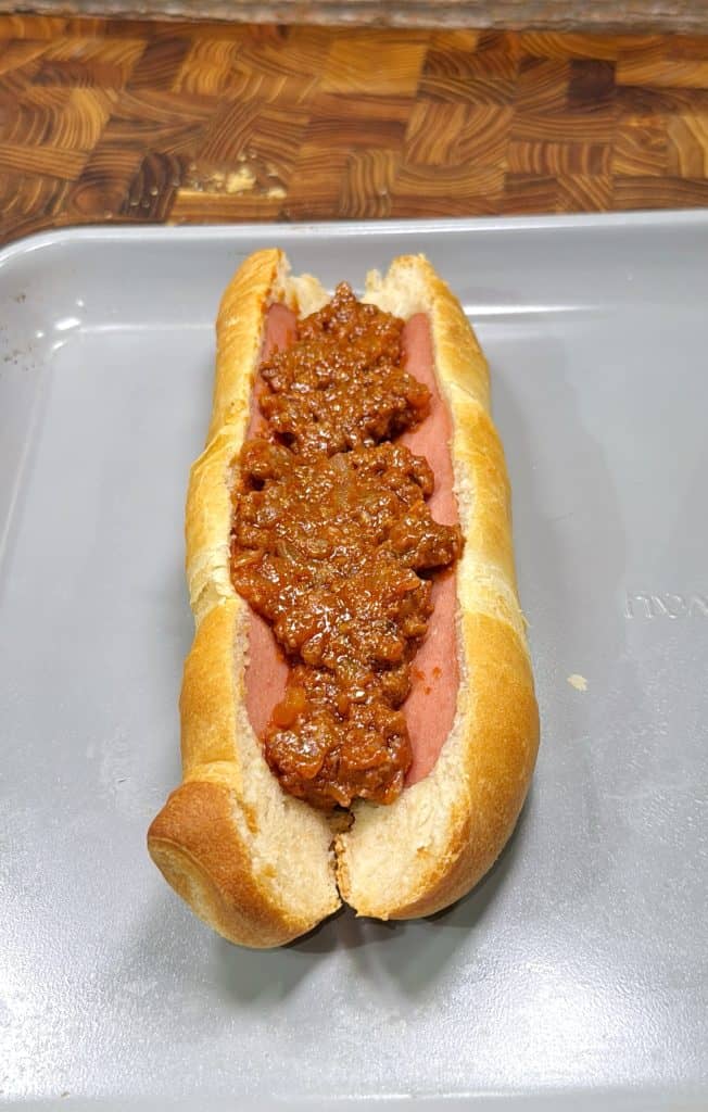 A hot dog in a bun topped with chili sits on a metal baking sheet, with a wooden cutting board in the background.