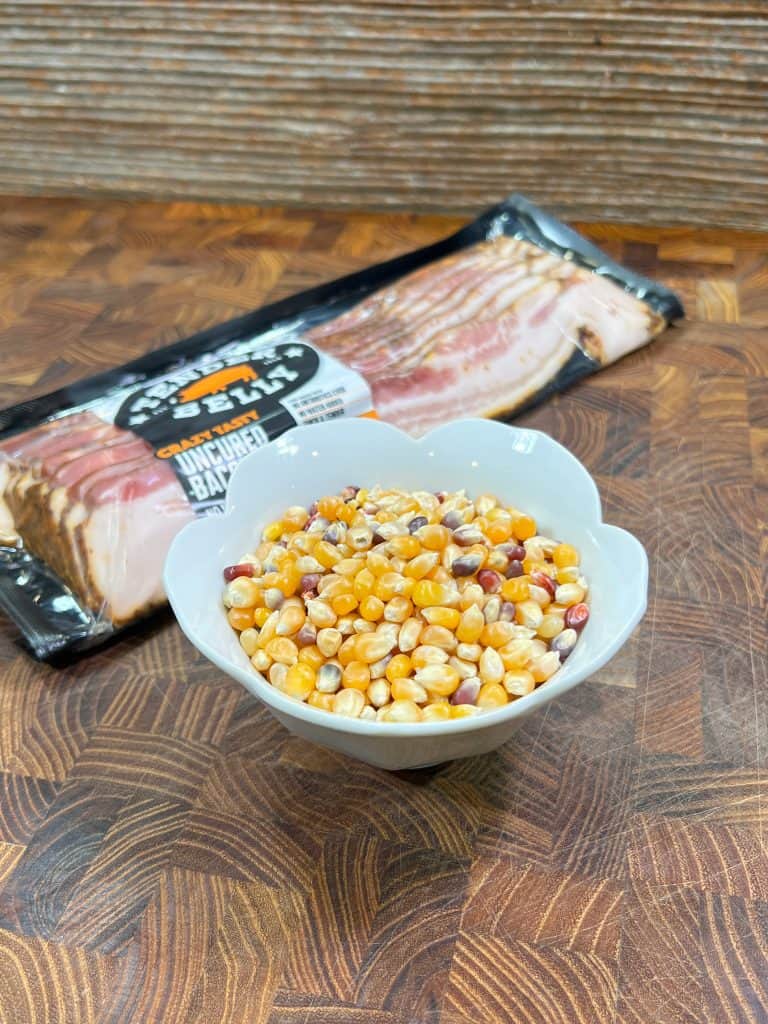 A white bowl filled with colorful popcorn kernels sits on a wooden surface, with a package of uncooked bacon placed behind it.