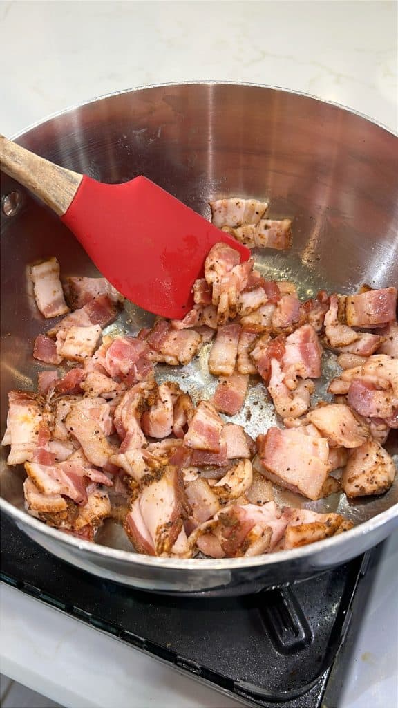 Diced bacon pieces being cooked in a stainless steel pan with a red silicone spatula on a stove.