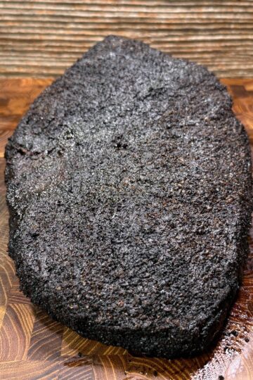 A large, dark, crusted smoked brisket rests on a wooden cutting board with a patterned surface and a wood-paneled background.