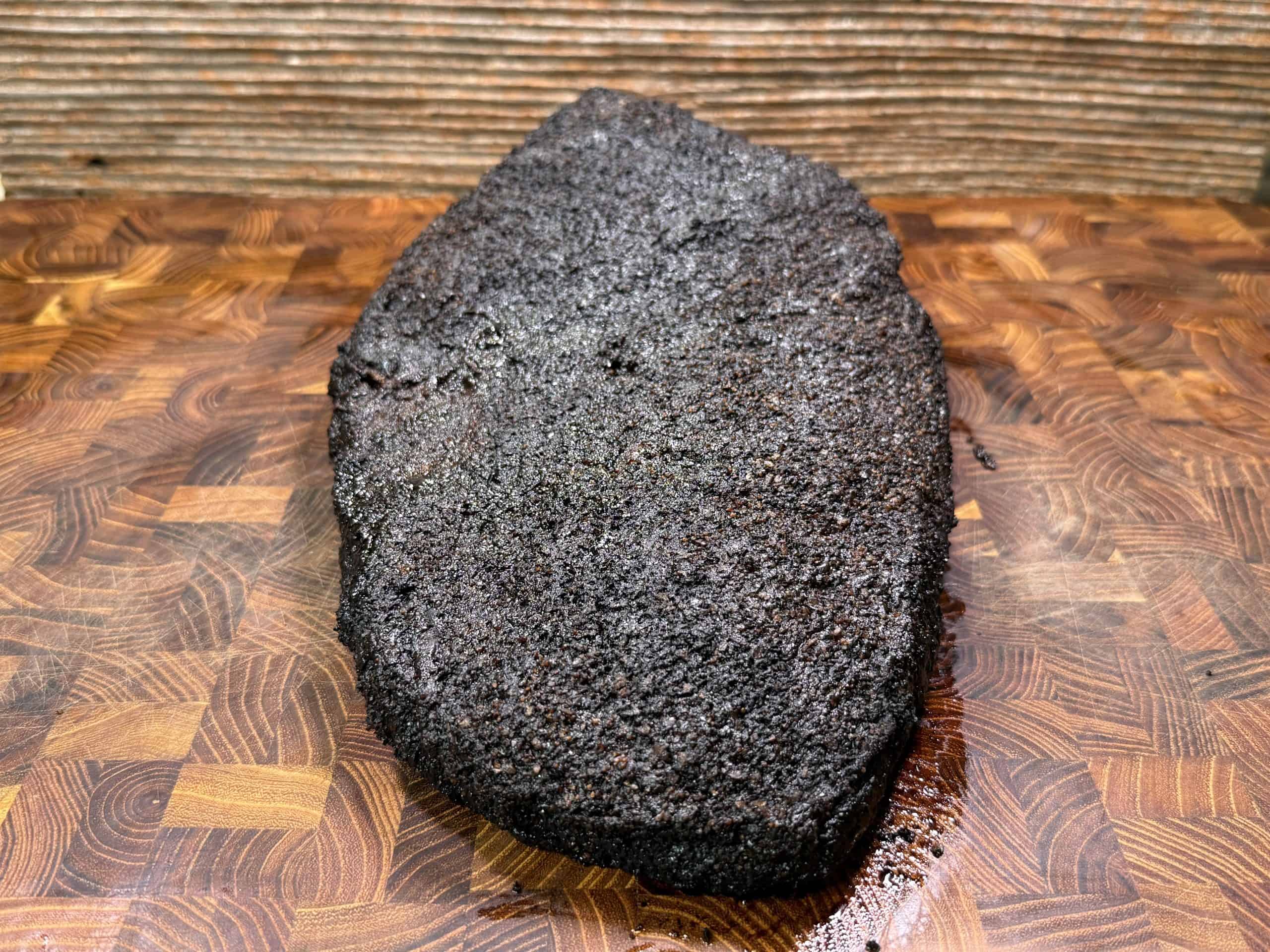 A large, dark, crusted smoked brisket rests on a wooden cutting board with a patterned surface and a wood-paneled background.