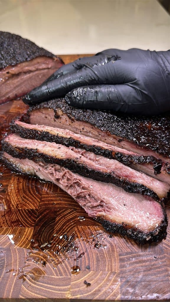 A gloved hand holds a smoked brisket on a wooden cutting board. Several thick, juicy slices with a dark bark and visible smoke ring are arranged in front, showcasing the meats texture and moisture.