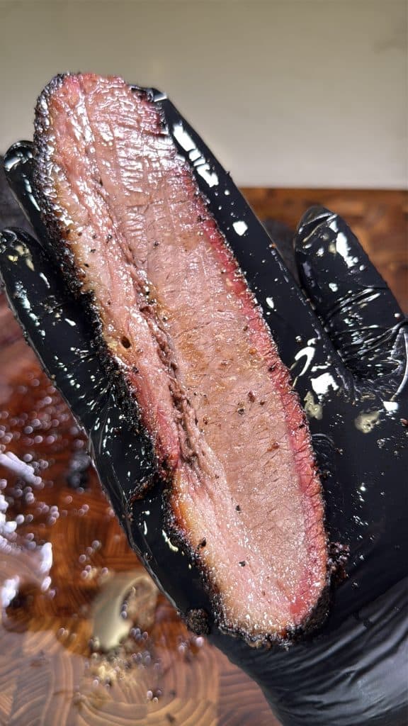 A gloved hand holds a thick, juicy slice of smoked no wrap brisket showing a barked crust, pink smoke ring, and tender, moist interior.