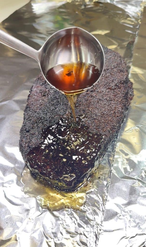 A ladle pours golden-brown liquid over a blackened, seasoned brisket resting on a sheet of aluminum foil.