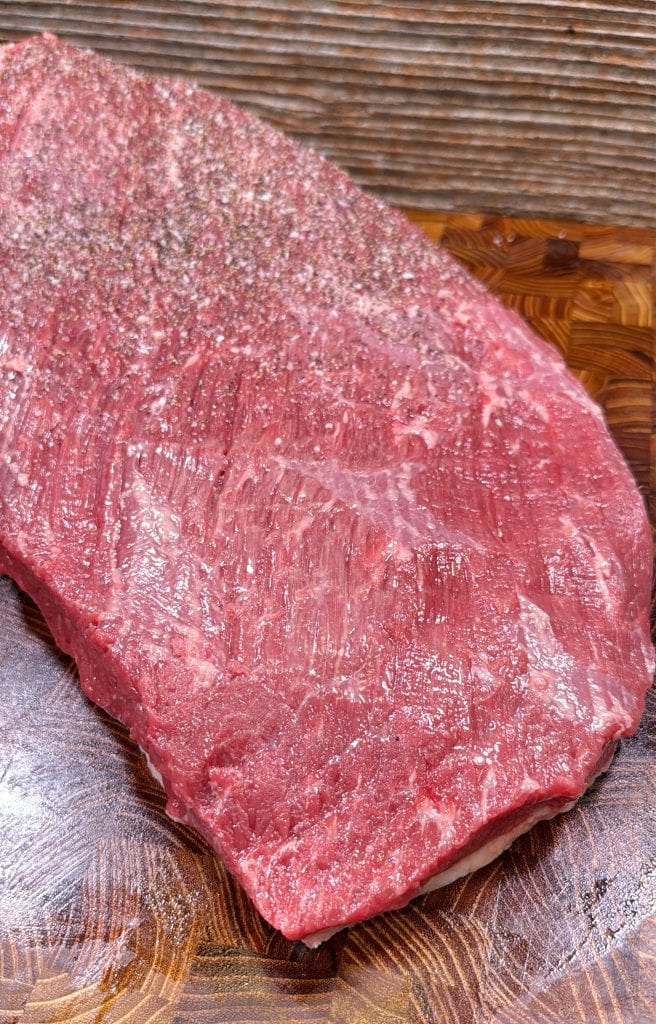 A close-up of a raw, seasoned piece of beef brisket resting on a wooden cutting board, with visible marbling and coarse black pepper sprinkled on top.