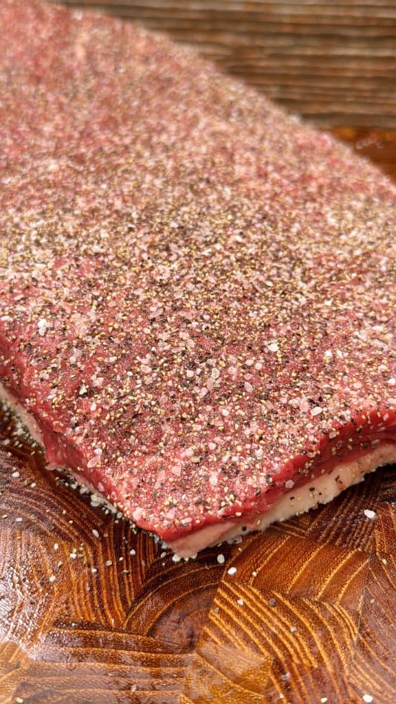 A raw slab of beef on a wooden surface, generously seasoned with coarse salt and cracked black pepper.