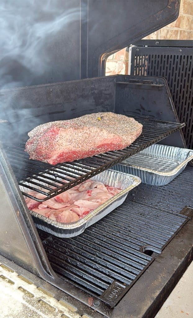 A large seasoned brisket sits on the top rack of a smoker, with two aluminum trays filled with liquid and sliced onions on the lower rack. Smoke is rising inside the smoker.