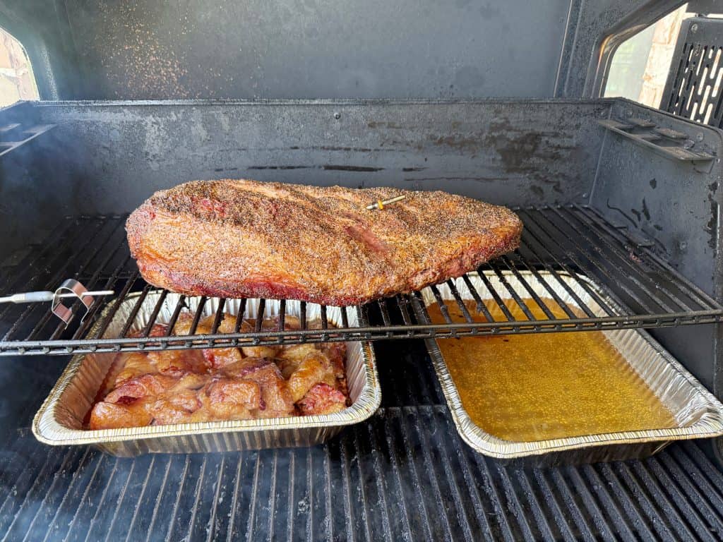 A large, seasoned brisket is smoking on the top rack of a grill. Below, aluminum trays contain meat and a yellowish liquid, likely for basting or moisture, with smoke rising inside the grill.
