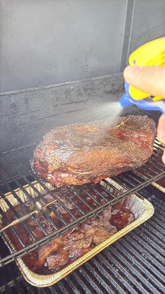 A hand uses a yellow spray bottle to mist a large, seasoned brisket on a grill rack, with a foil tray catching drippings below inside a smoker.
