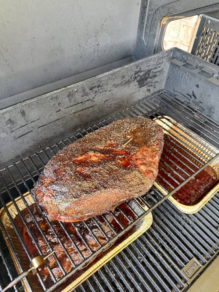 A large, seasoned brisket cooks on the rack of a smoker, with a pan underneath catching drippings. The meat has a dark, crispy bark forming on its surface.